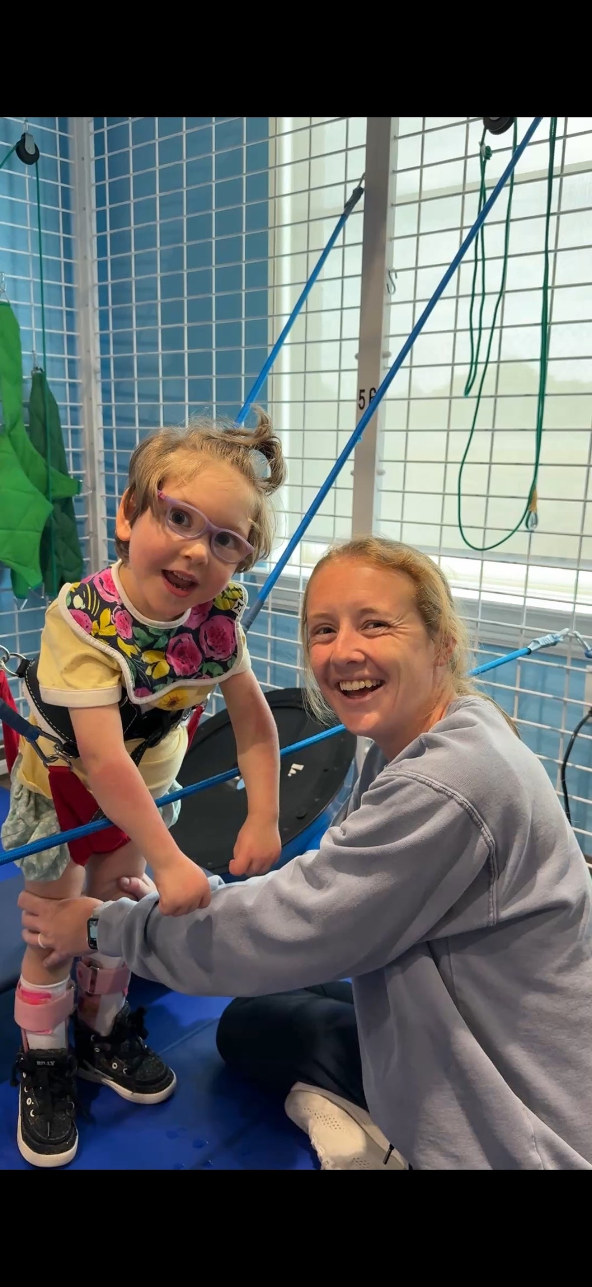 Gracie standing in supportive therapy equipment beside a smiling adult.