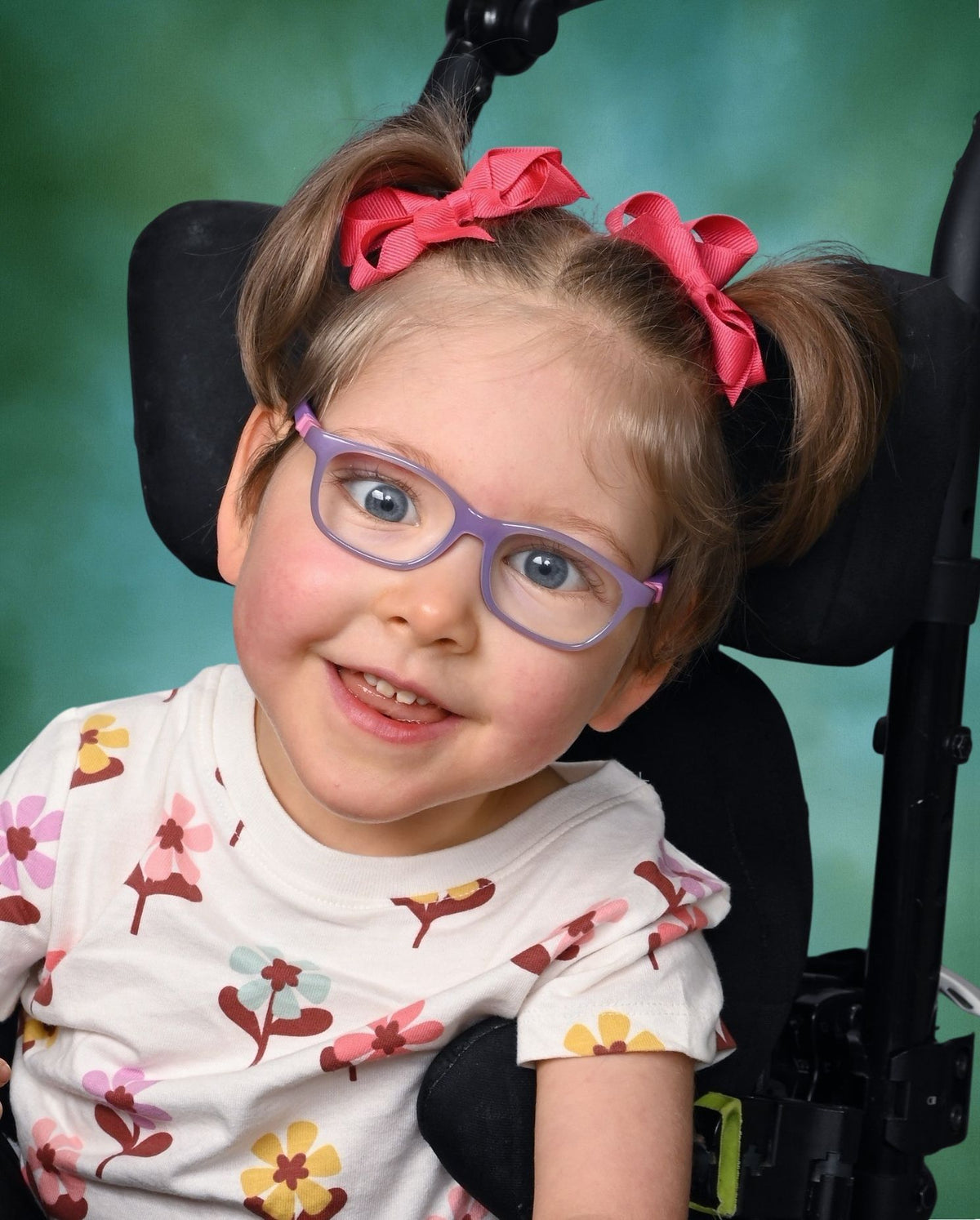 Gracie smiling in purple glasses and pink hair bows against a green background.