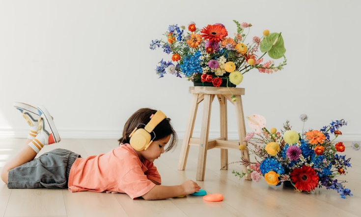 A child lying on the floor with headphones beside flowers on a stool